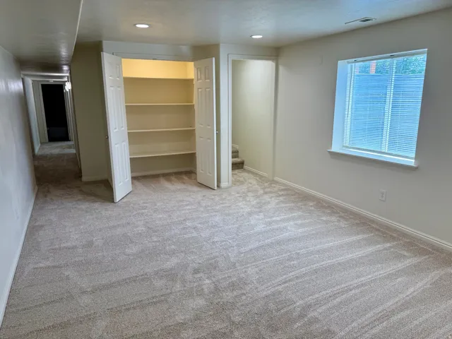 Spacious carpeted room featuring a built-in closet with shelving, a window with blinds, recessed lighting, and a doorway to a hallway