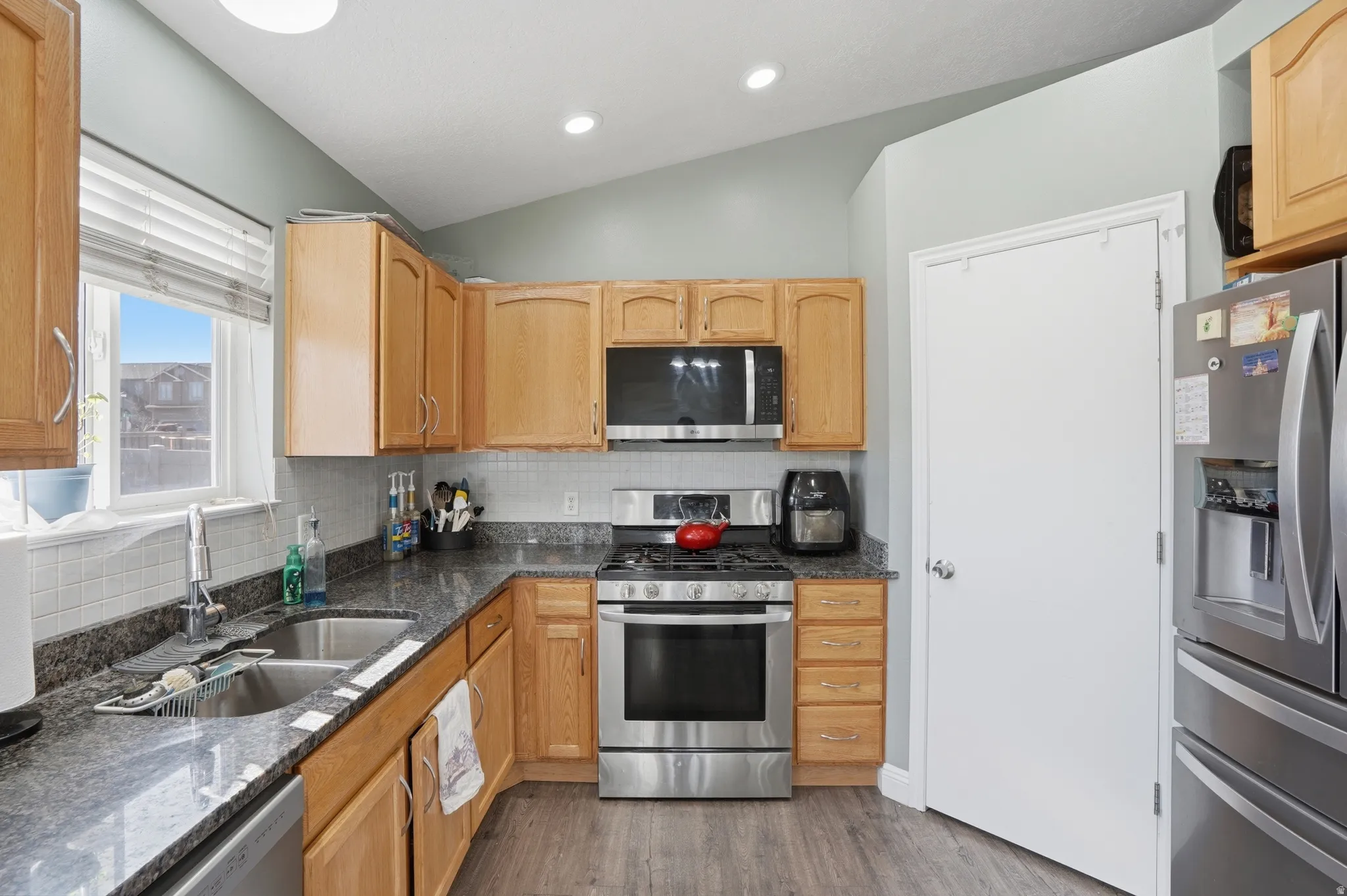 Kitchen featuring stainless steel appliances, vaulted ceiling, dark stone counters, decorative backsplash, and light wood finish cabinets