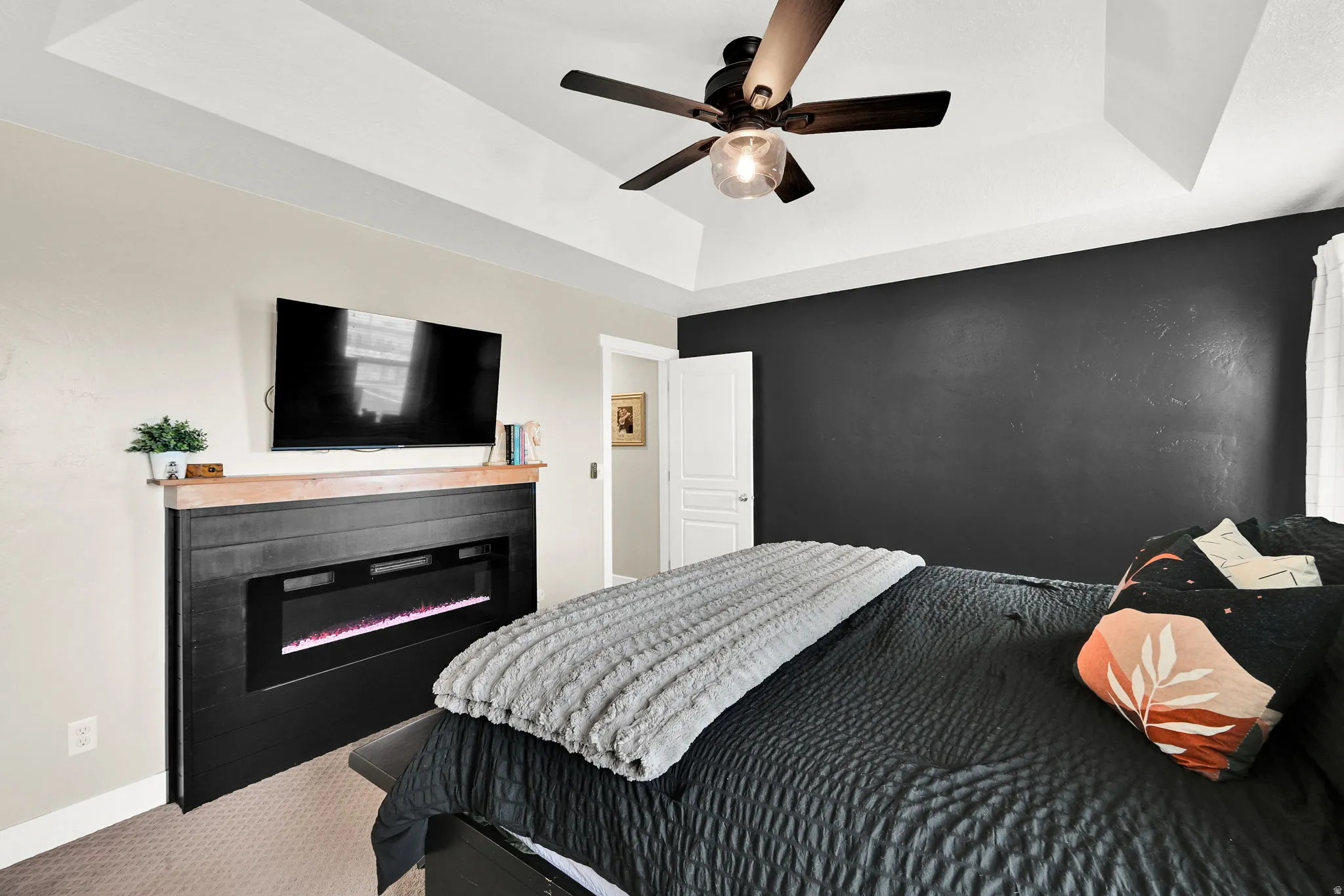 Carpeted bedroom with a tray ceiling, ceiling fan, and a glass covered fireplace