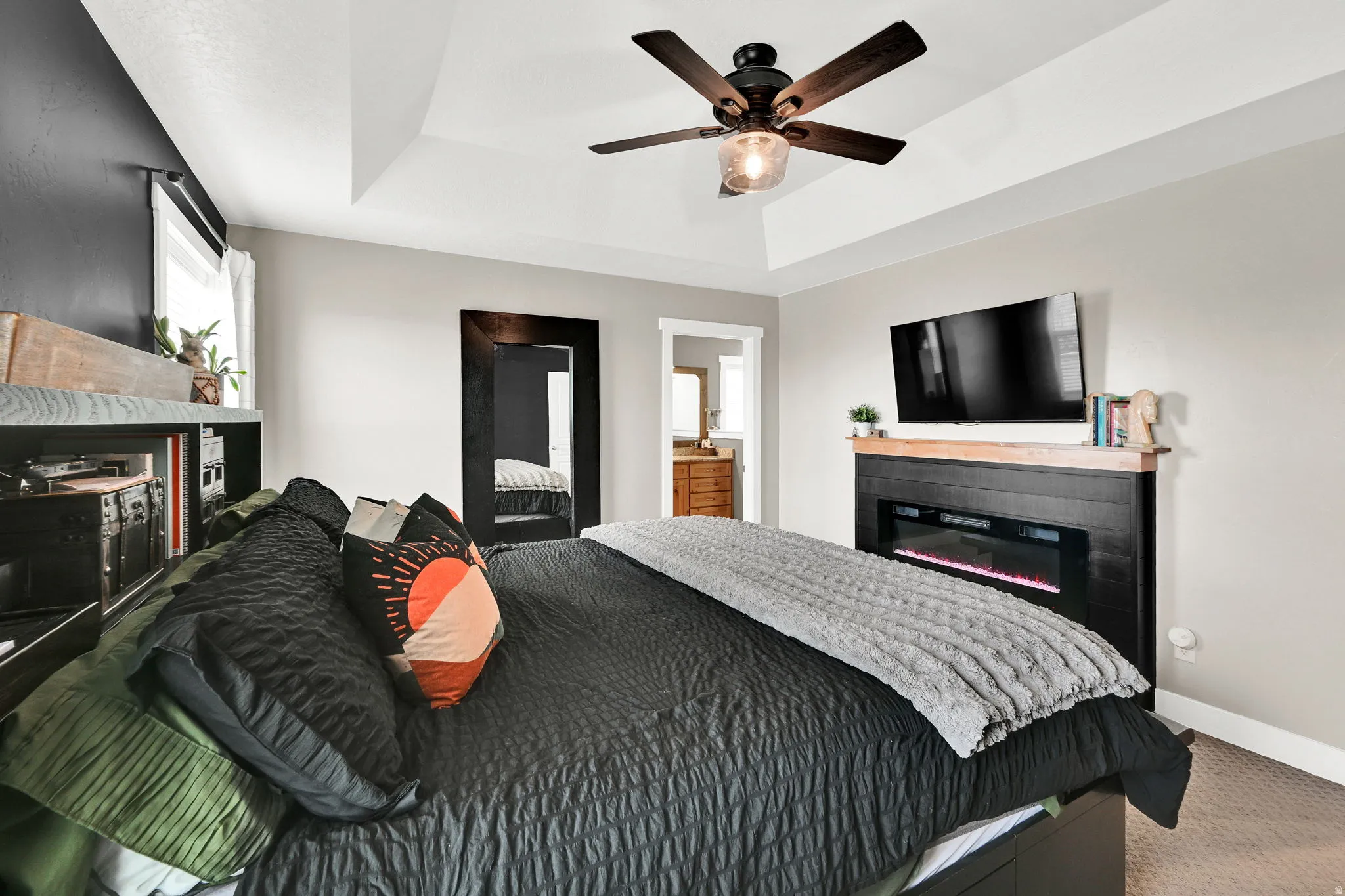 Carpeted bedroom featuring a glass covered fireplace, a tray ceiling, a ceiling fan, and connected bathroom