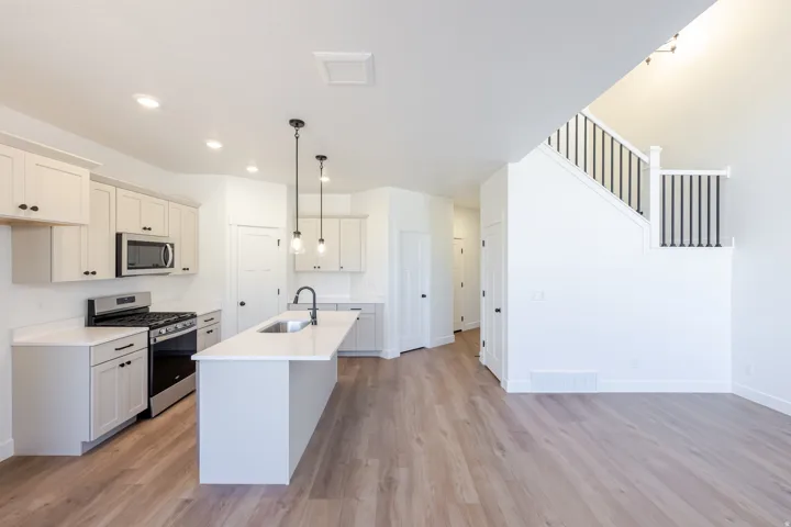 Open-concept kitchen and living area featuring wood-finish flooring, white cabinetry, a kitchen island with sink, stainless steel appliances, and a staircase with black metal balusters