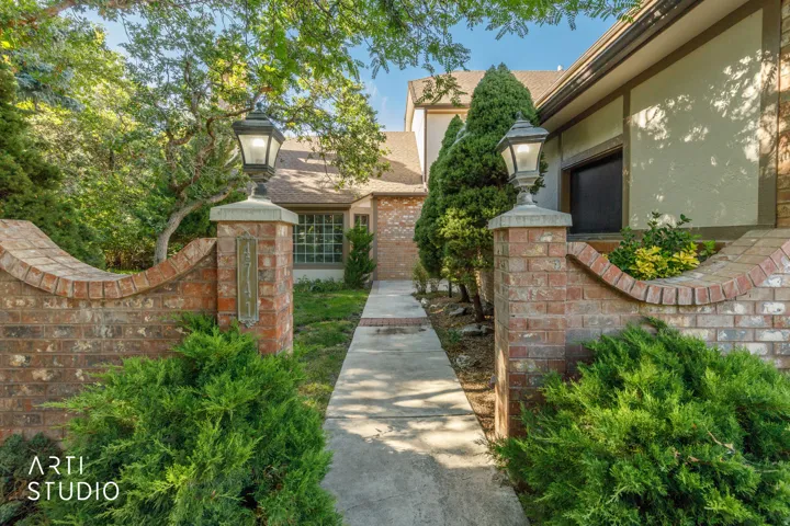Brick wall with integrated lighting fixtures, mature landscaping, and a concrete pathway leading to the residence