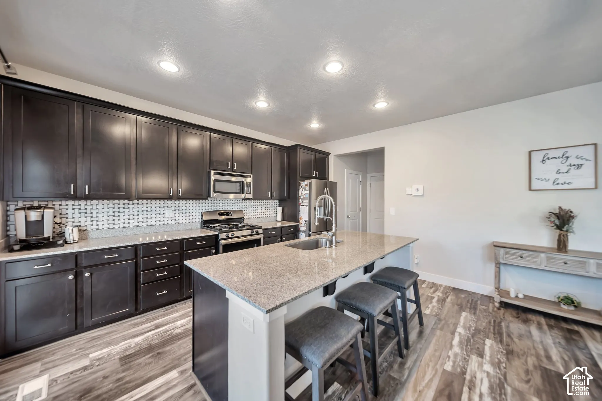 Island Kitchen with stainless steel appliances, light stone counters, light wood-style floors, and an island with sink