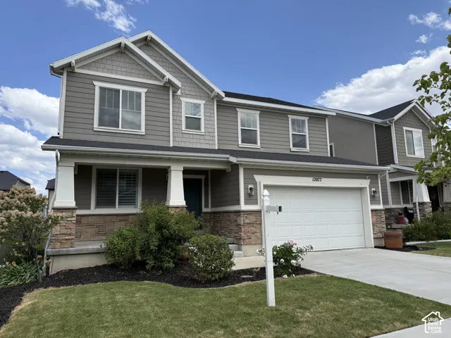 Two-story home featuring a combination of horizontal siding and shake siding accents, a covered front porch with stone base columns, an attached garage with a concrete driveway, and a manicured lawn with landscape beds