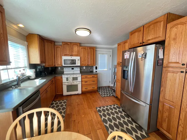 Kitchen with wood finish cabinetry, stainless steel and white appliances, dark countertops, light wood-style floors, and crown molding