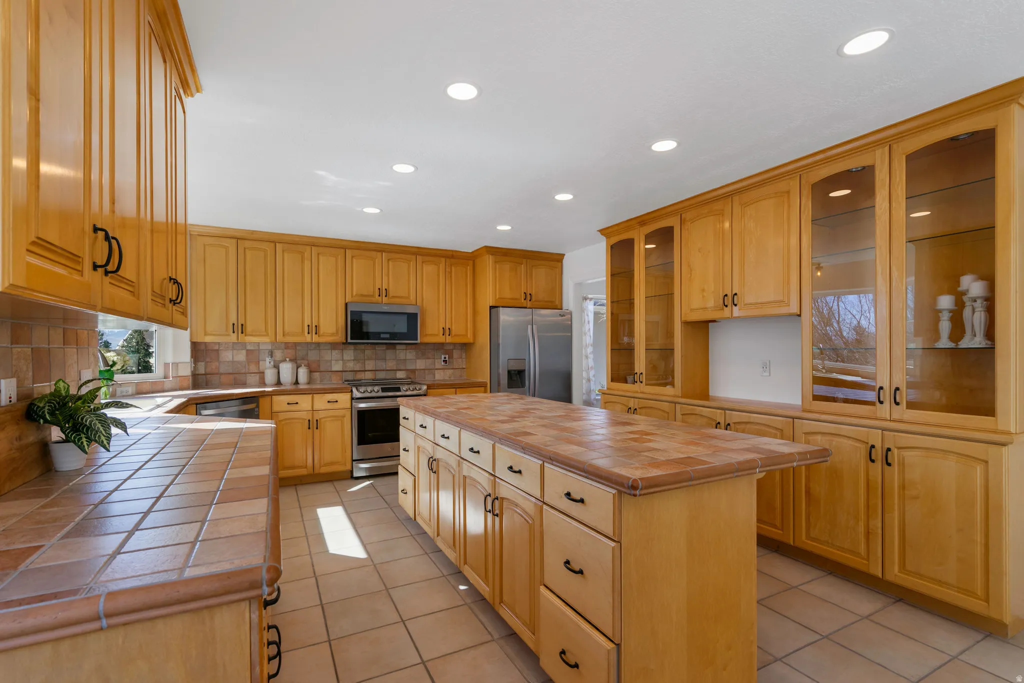 Kitchen with tile countertops, stainless steel appliances, glass fronted cabinets, a center island, and recessed lighting