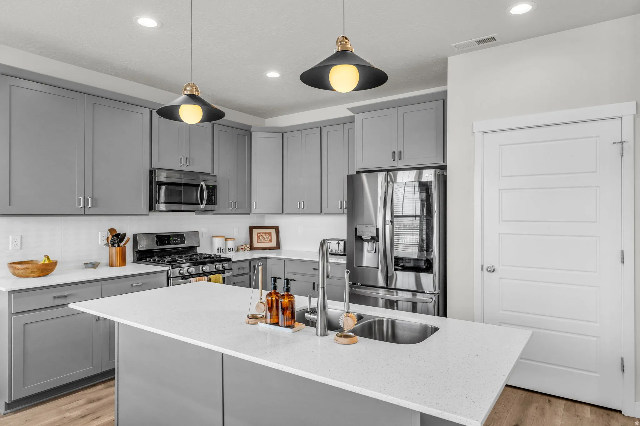 Kitchen featuring gray cabinets, stainless steel appliances, a kitchen island with sink, hanging light fixtures, and light wood-style flooring