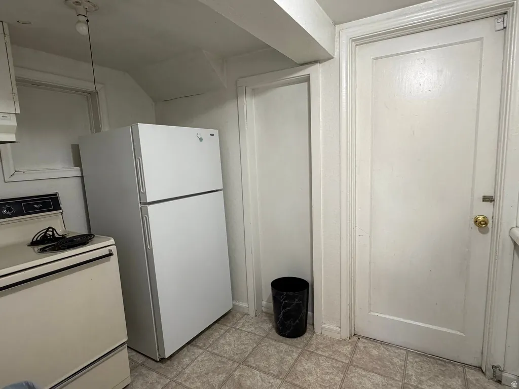 White paneled interior door, utility door frame, overhead bare bulb light fixture, white refrigerator, and tiled flooring