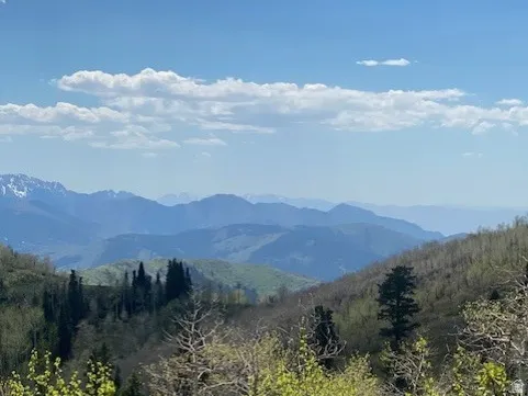 Expansive mountain landscape featuring distant peaks, verdant hillsides, and a clear blue sky with scattered clouds