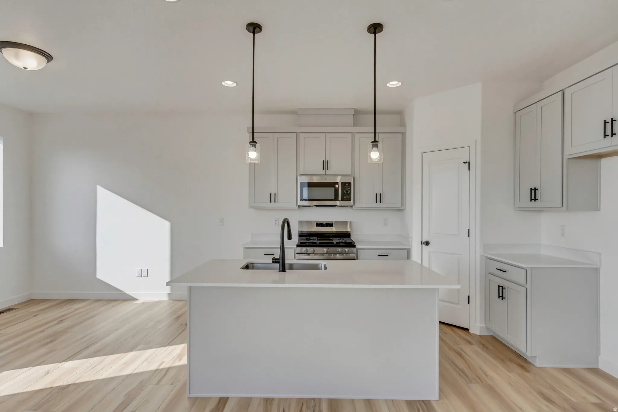Kitchen with stainless steel appliances, light wood-style floors, a kitchen island with sink, decorative light fixtures, and light stone countertops