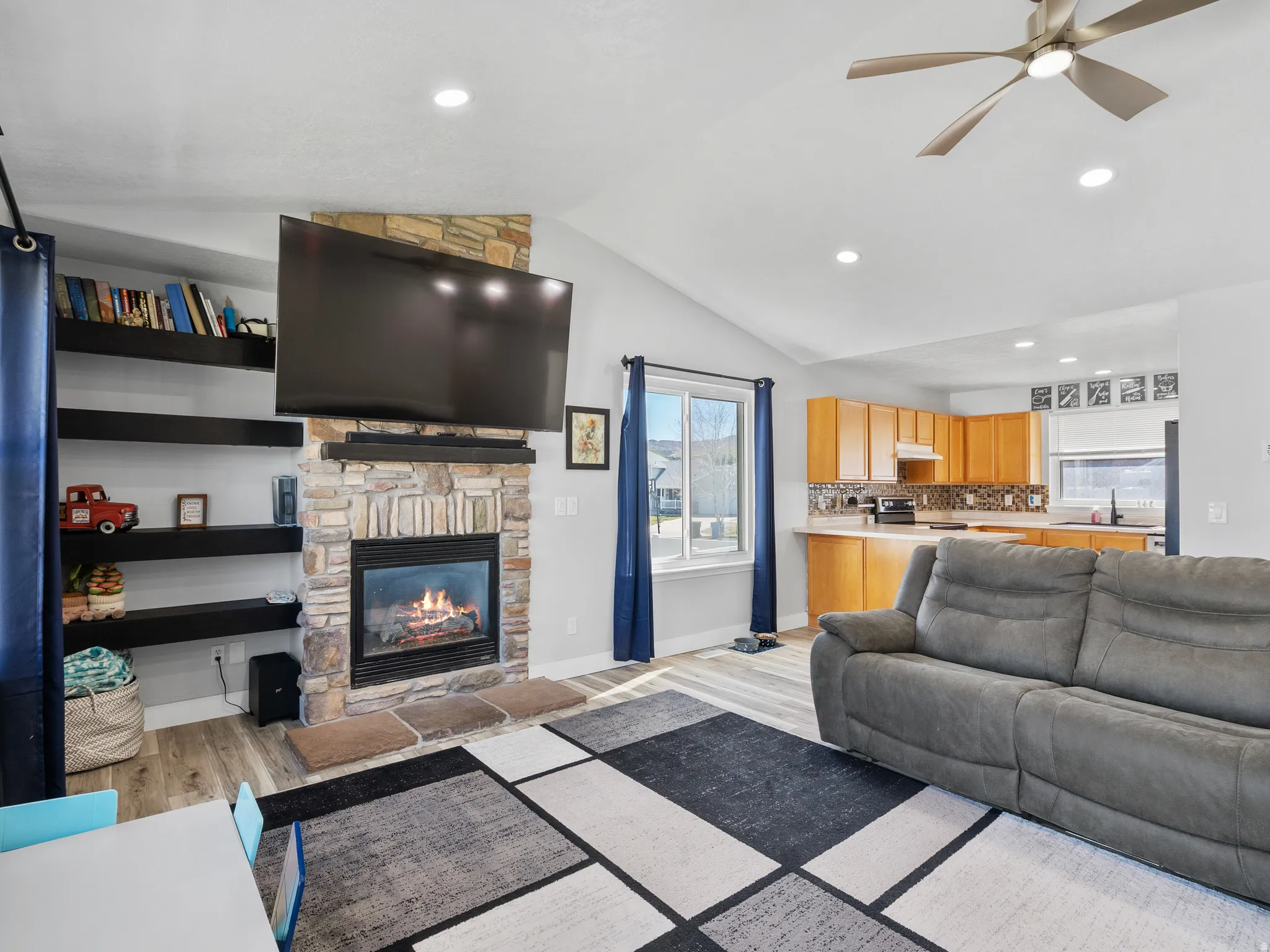 Living room featuring light wood-style flooring, a ceiling fan, vaulted ceiling, a stone fireplace, and recessed lighting
