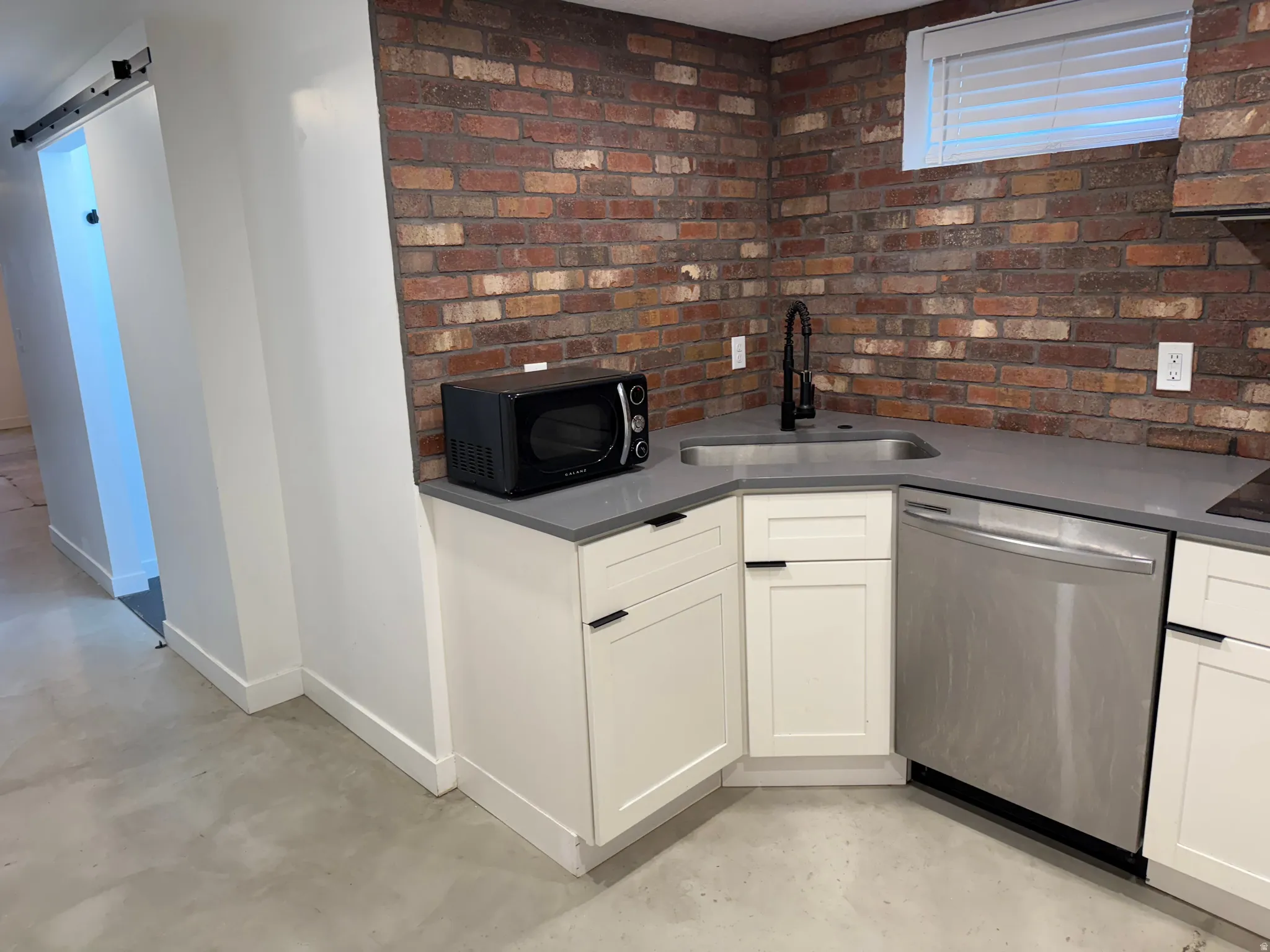 Kitchen with dishwasher, concrete flooring, white cabinetry, black microwave, and brick wall