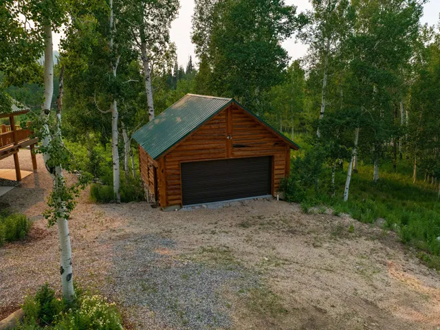 Log cabin style detached garage with a dark green metal roof and a dark brown overhead door, surrounded by mature trees and natural landscaping