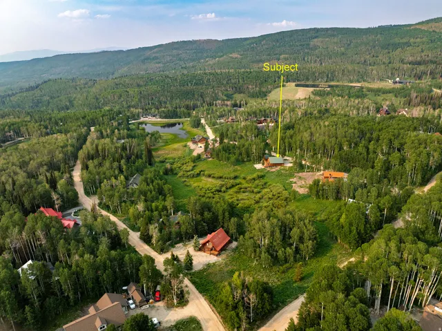 Aerial perspective showcasing a single-story dwelling surrounded by dense forest, with a nearby pond and mountainous terrain in the background