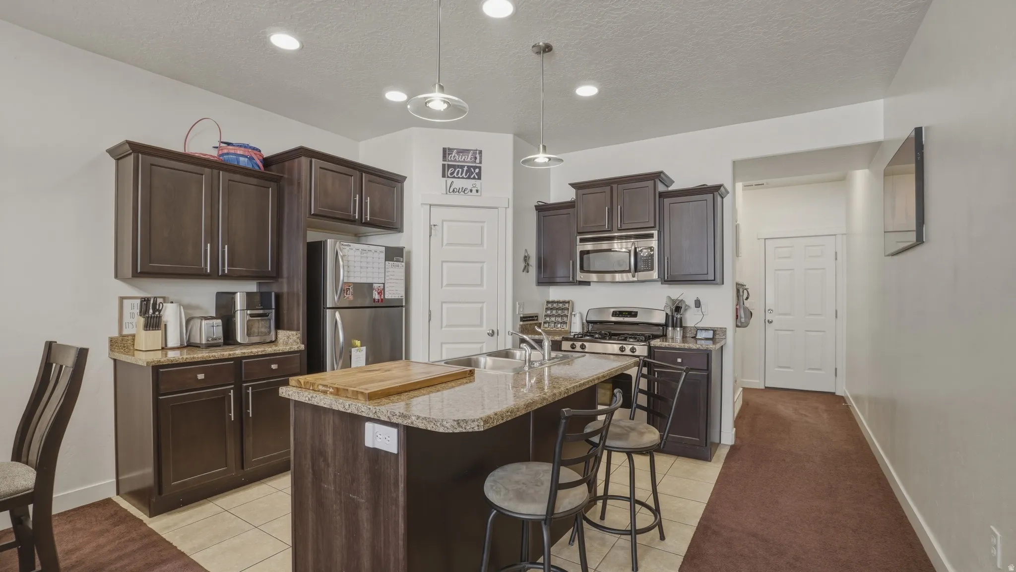 Kitchen featuring dark wood finish cabinets, stainless steel appliances, a kitchen bar, a kitchen island with sink, and pendant lighting