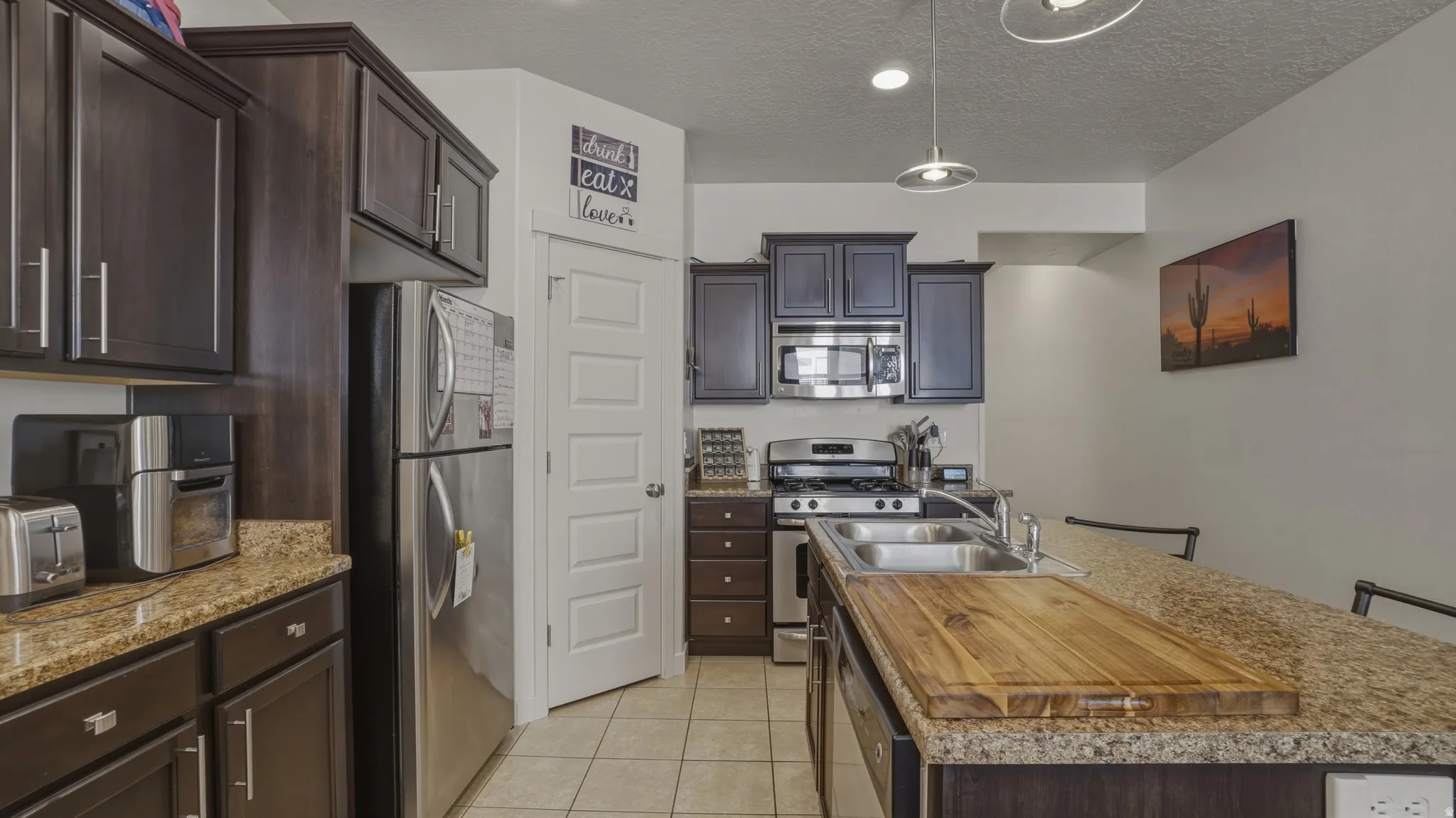 Kitchen with a kitchen island with sink, dark wood finish cabinetry, stainless steel appliances, light tile patterned floors, and decorative light fixtures