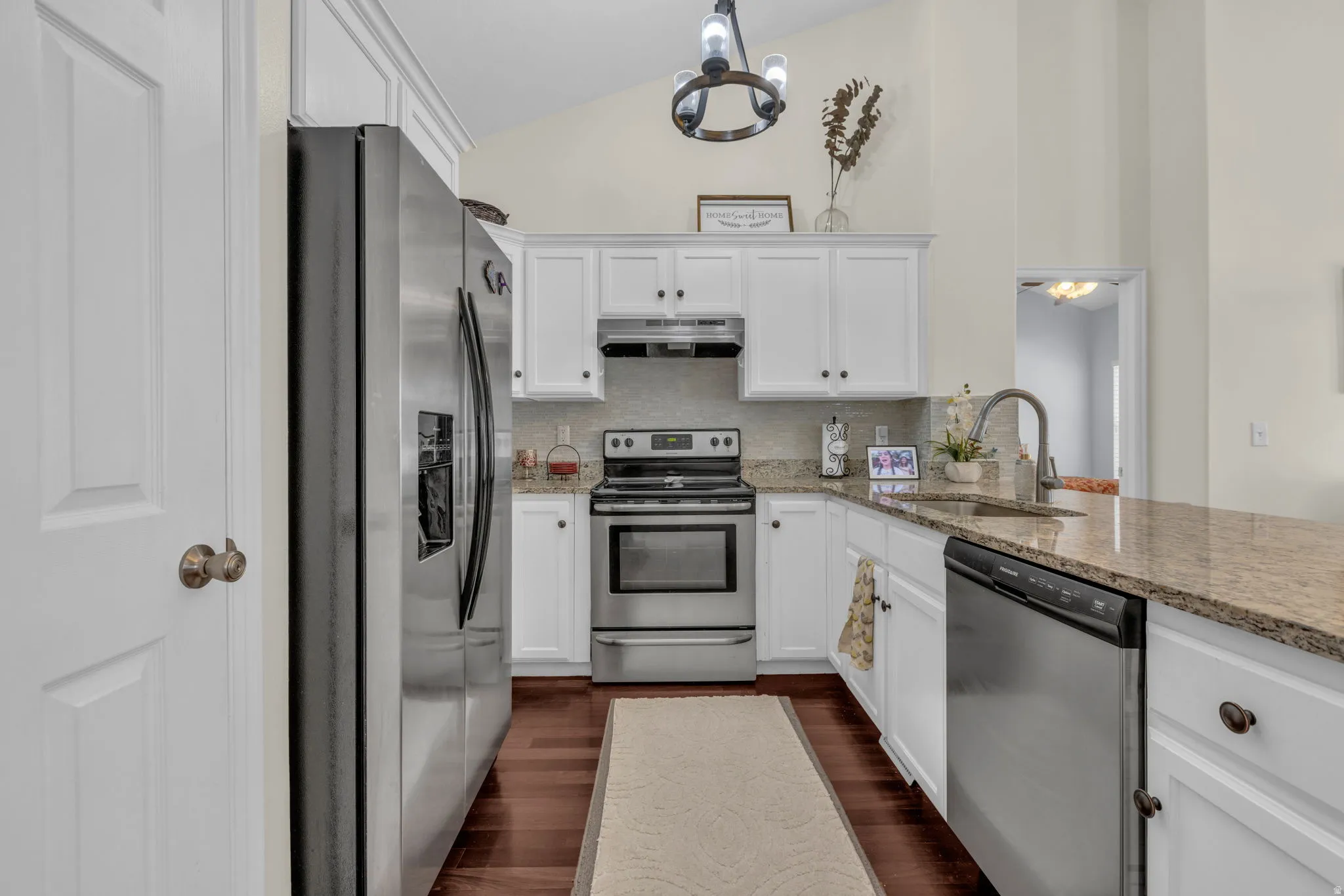 Kitchen with stainless steel appliances, lofted ceiling, white cabinetry, light stone countertops, and dark wood finished floors