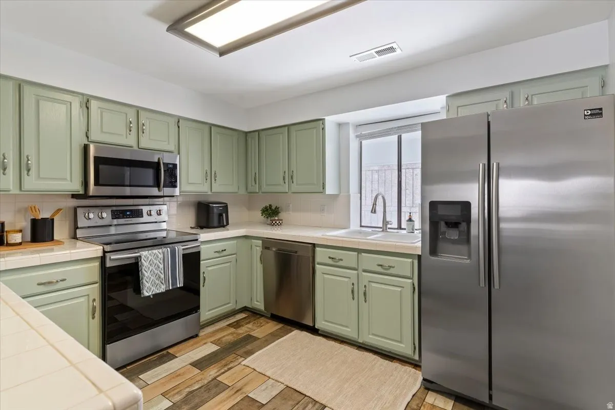 Kitchen featuring tile countertops, green cabinetry, stainless steel appliances, light wood-style floors, and backsplash