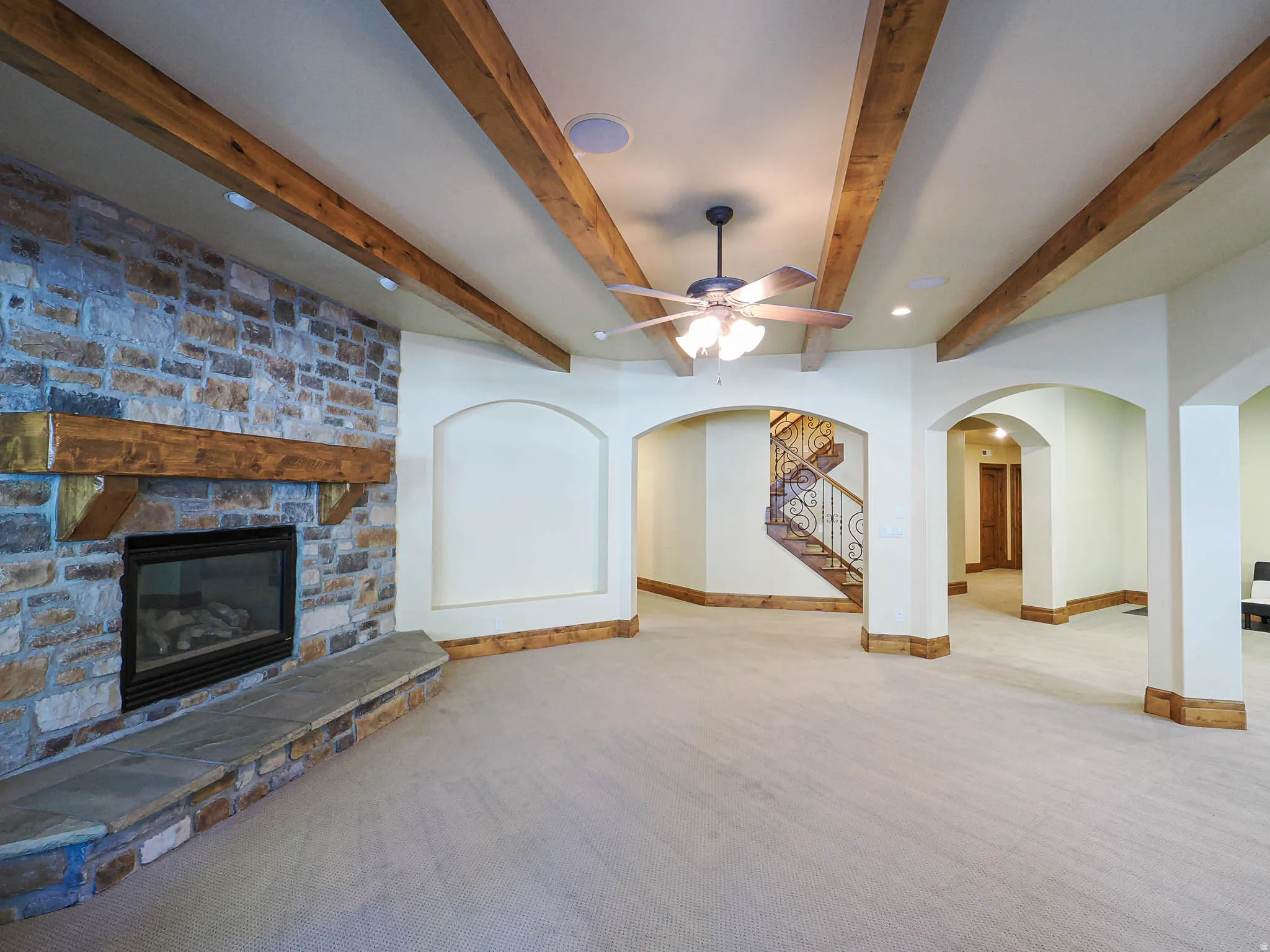 Unfurnished living room with arched walkways, beamed ceiling, a stone fireplace, ceiling fan, and carpet