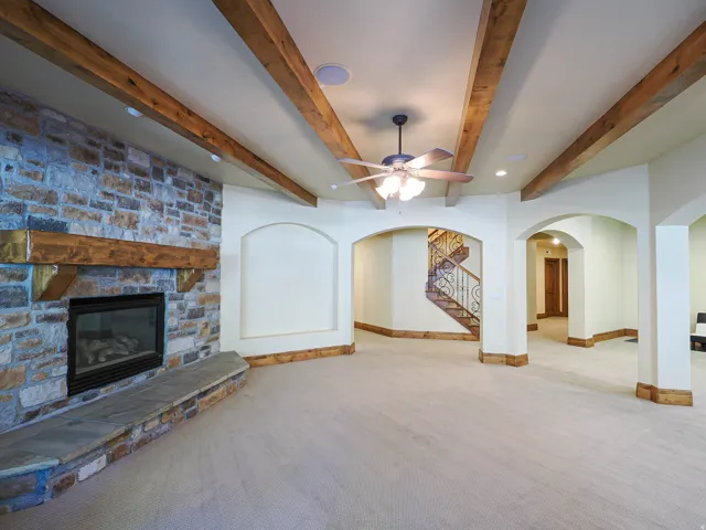 Unfurnished living room with arched walkways, beamed ceiling, a stone fireplace, ceiling fan, and carpet