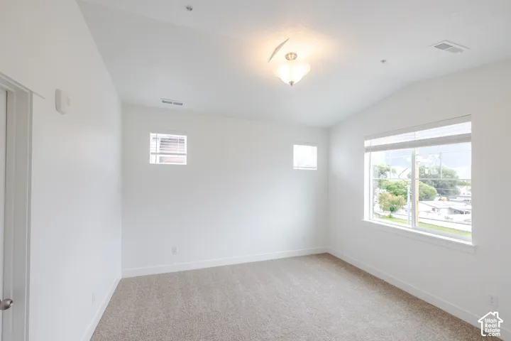 Neutral tone carpeting, vaulted ceiling, flush mount ceiling light fixture, large window with blinds, and additional window