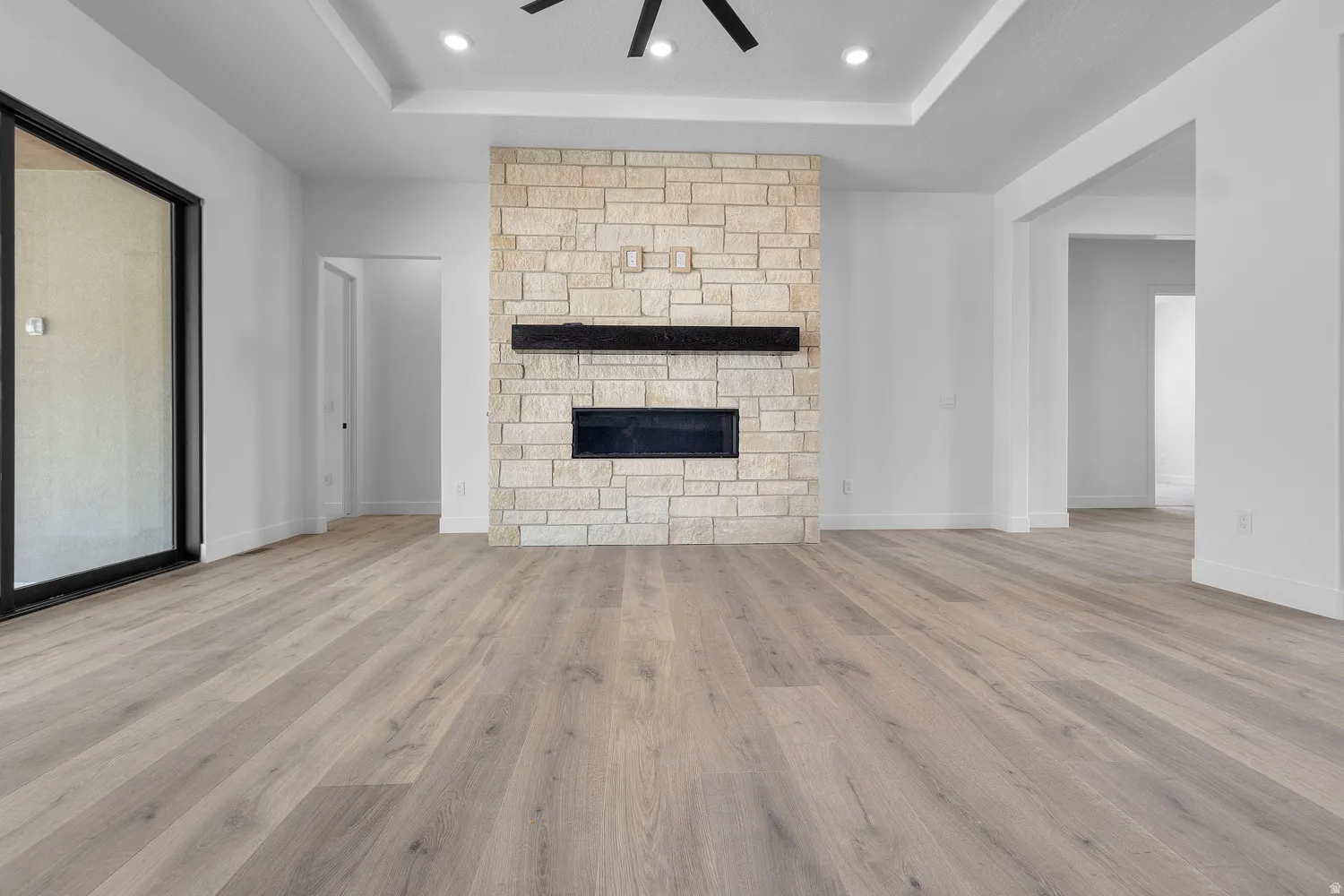 Unfurnished living room featuring light wood-type flooring, ceiling fan, a tray ceiling, a stone fireplace, and recessed lighting