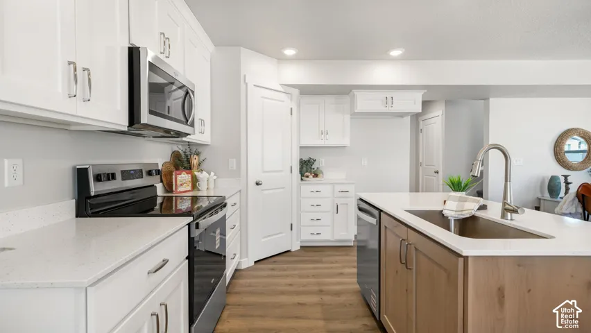 Contemporary kitchen featuring white cabinetry, stainless steel appliances, light-toned countertops, a kitchen island with a built-in sink, and wood-finish flooring