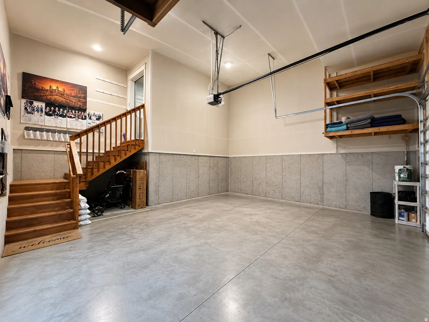 Spacious garage featuring polished concrete flooring, extensive wall-mounted shelving with wooden frames, exposed ceiling infrastructure with pendant lighting, and a wooden staircase with railings providing access to upper-level spaces