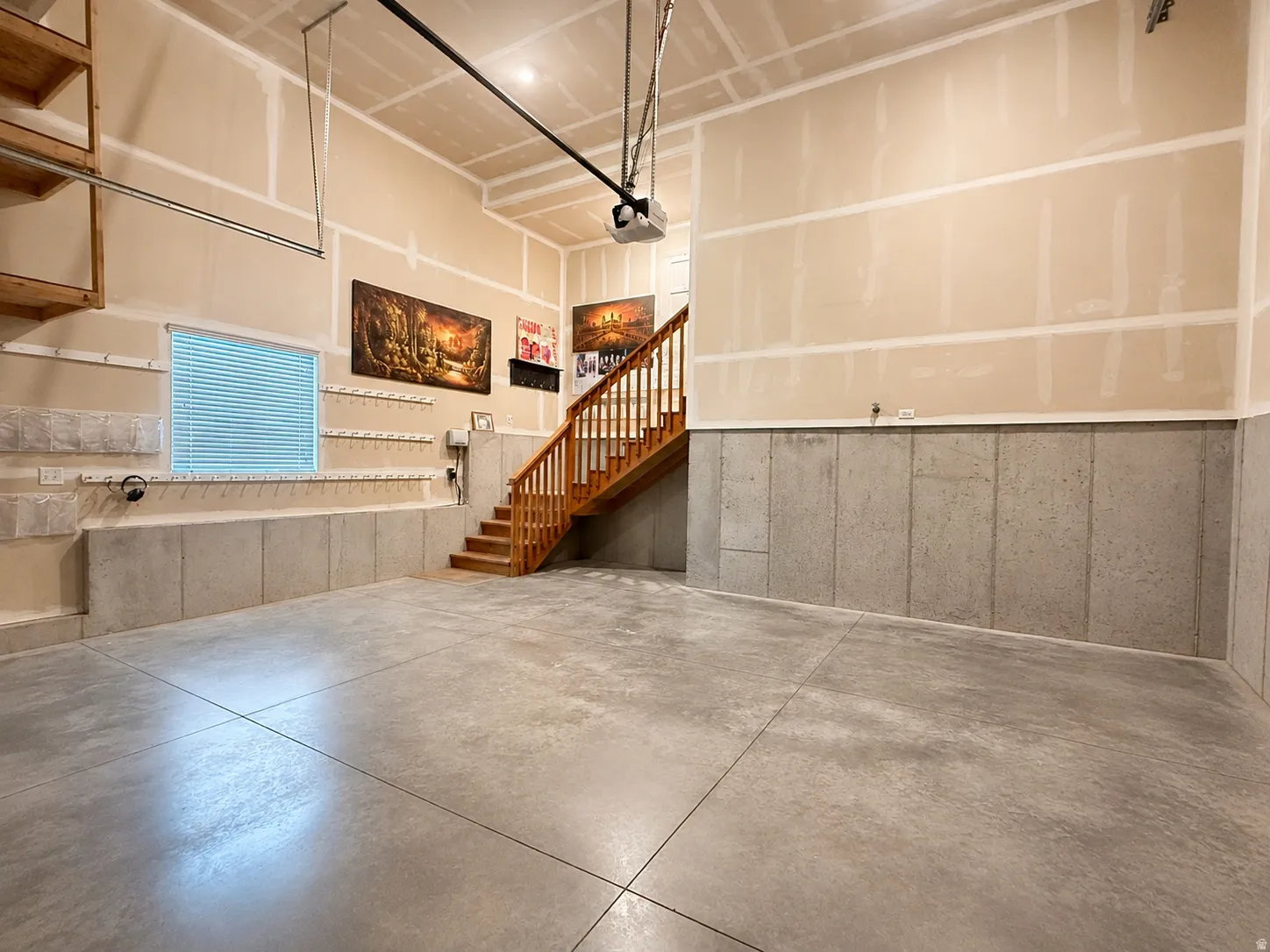 Concrete flooring, unfinished walls, built-in shelving, wood staircase with railing, and a window with blinds