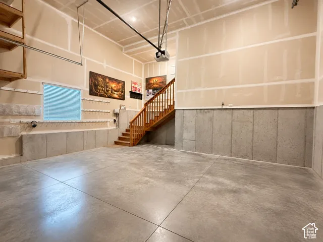 Concrete flooring, unfinished walls, built-in shelving, wood staircase with railing, and a window with blinds