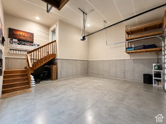 Spacious garage featuring polished concrete flooring, extensive wall-mounted shelving with wooden frames, exposed ceiling infrastructure with pendant lighting, and a wooden staircase with railings providing access to upper-level spaces