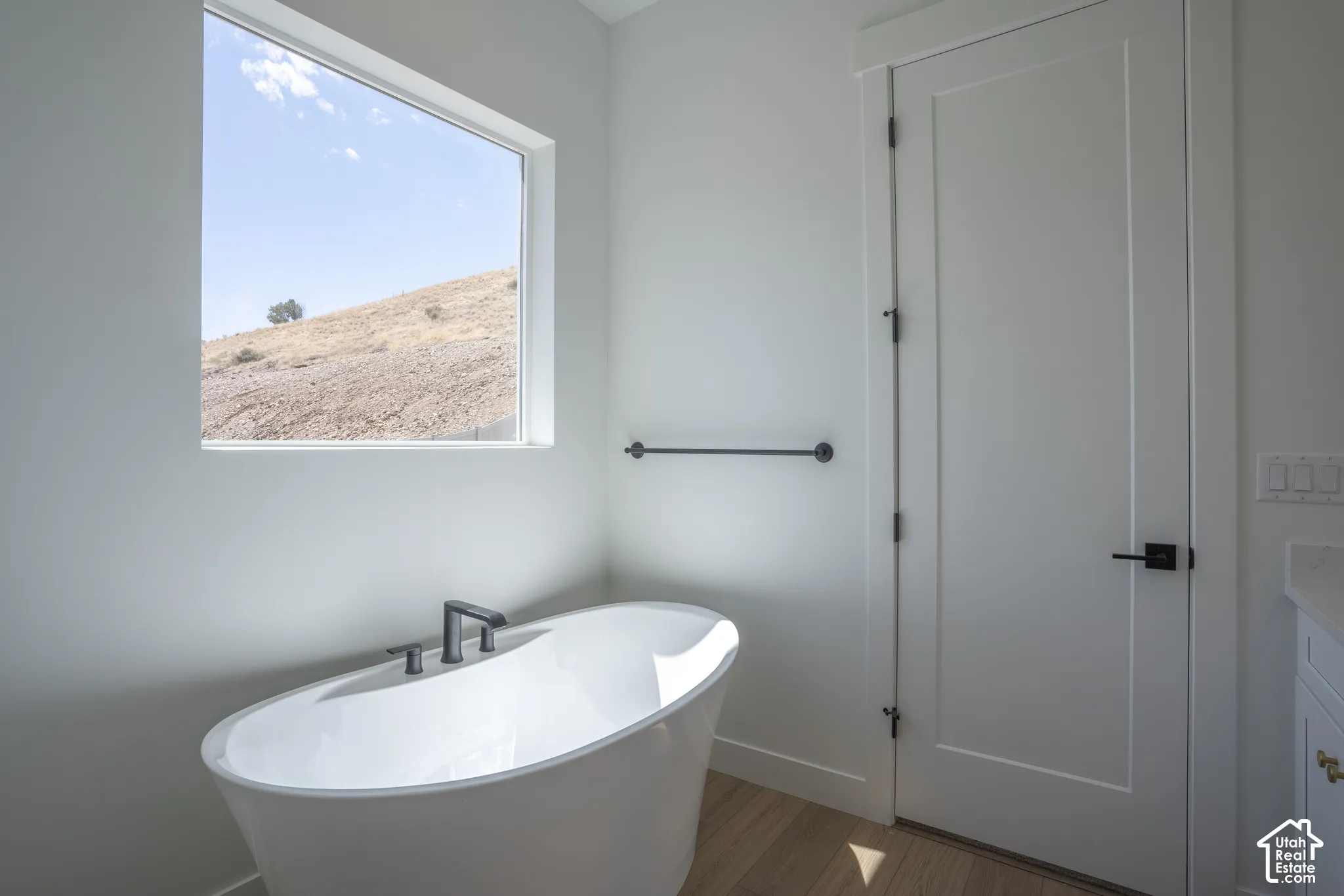 Freestanding soaking tub with dark-finish fixtures, a large picture window, light wood-finish flooring, and a solid core door with dark-finish hardware