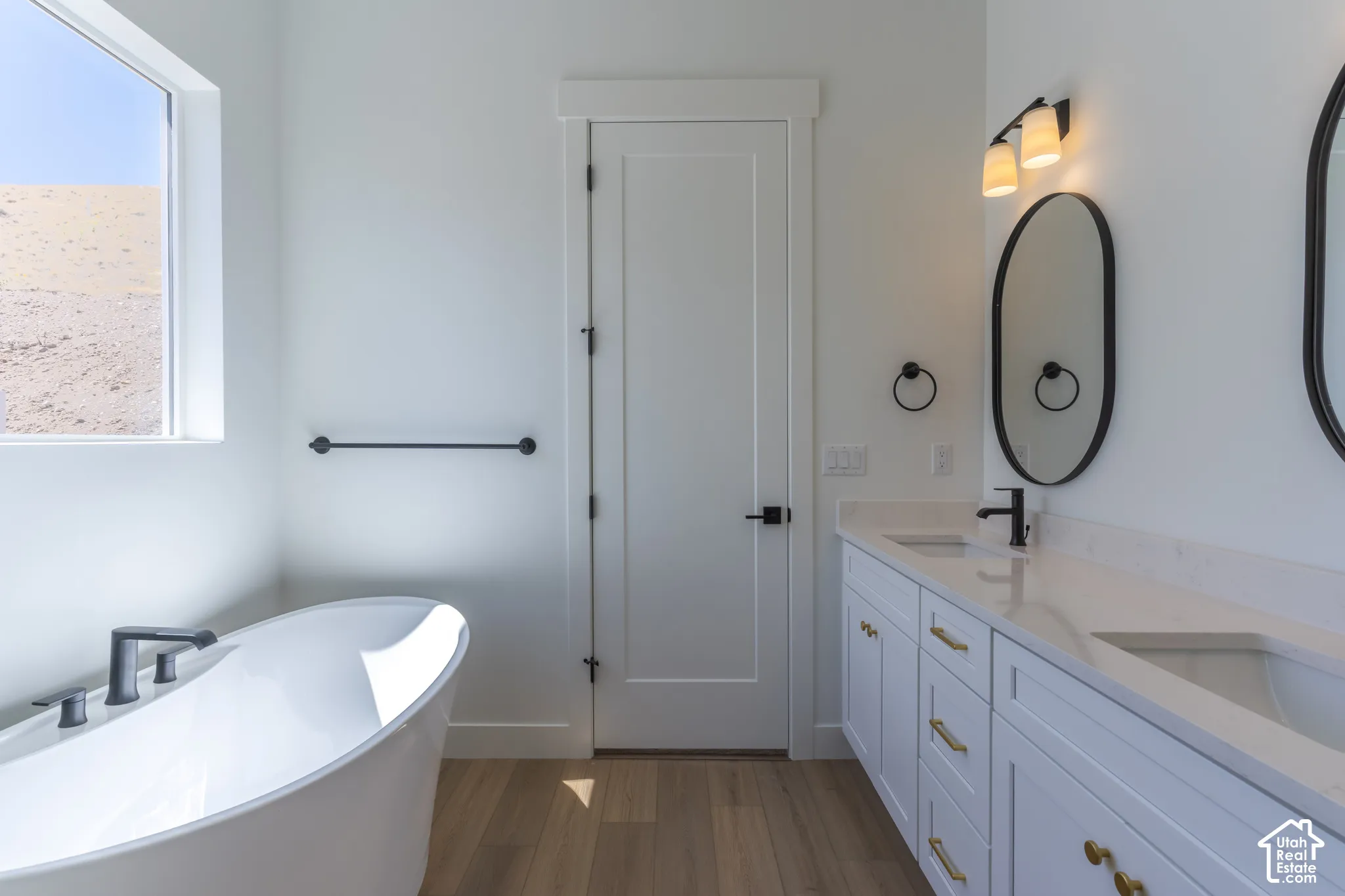 Freestanding soaking tub with a matte black faucet, dual vanity with white cabinetry, gold-finish hardware, and a stone-finish countertop, wood-finish flooring, and white walls