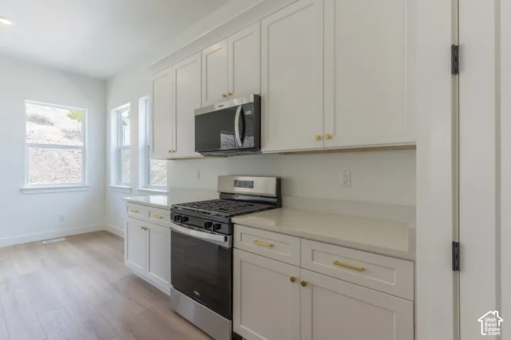 White cabinetry with gold-tone hardware, stainless steel appliances, light-toned countertops, wood-finish flooring, and multiple windows