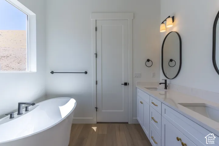 Freestanding soaking tub with a matte black faucet, dual vanity with white cabinetry, gold-finish hardware, and a stone-finish countertop, wood-finish flooring, and white walls