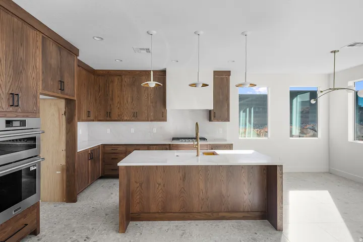 Kitchen featuring a center island with sink, light stone counters, stainless steel appliances, backsplash, and wood finish cabinetry