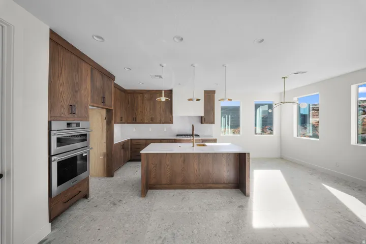 Kitchen featuring an island with sink, light stone countertops, double oven, decorative light fixtures, and light floors