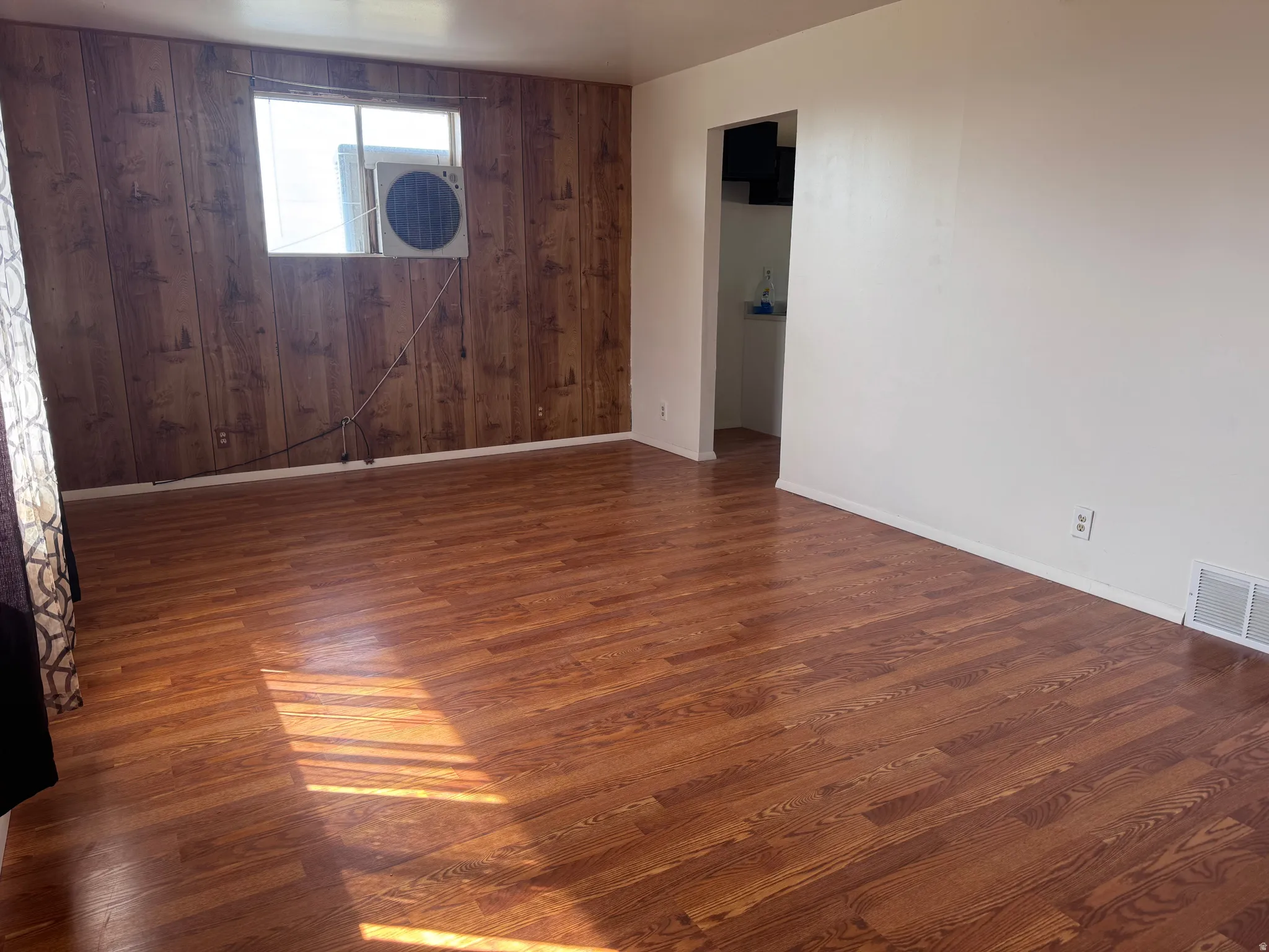 Spacious room featuring wood-finish flooring, a paneled accent wall, and a window with an integrated air conditioning unit
