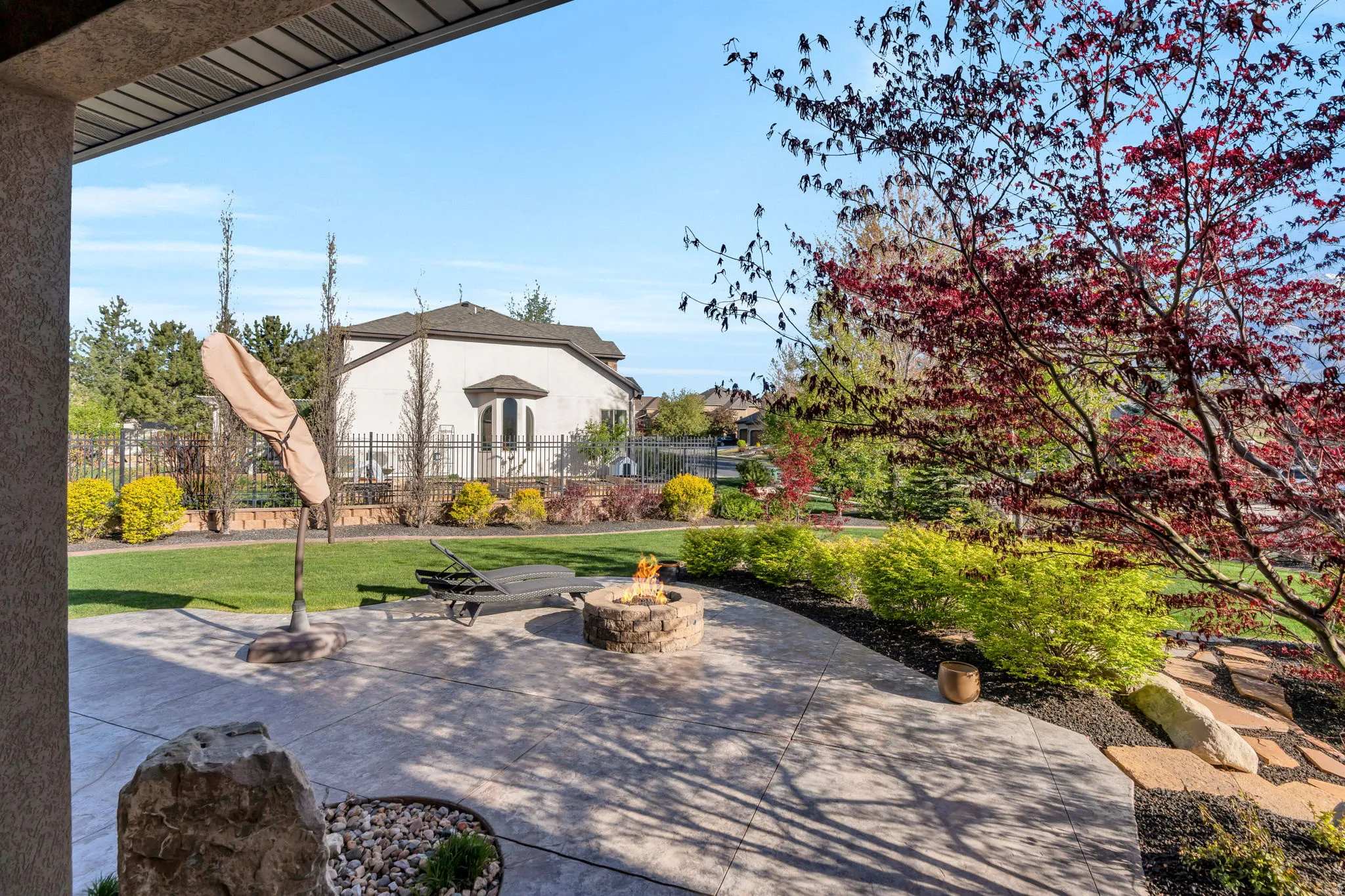 Expansive stamped concrete patio featuring a built-in fire pit, surrounded by lush landscaping with mature trees and shrubs