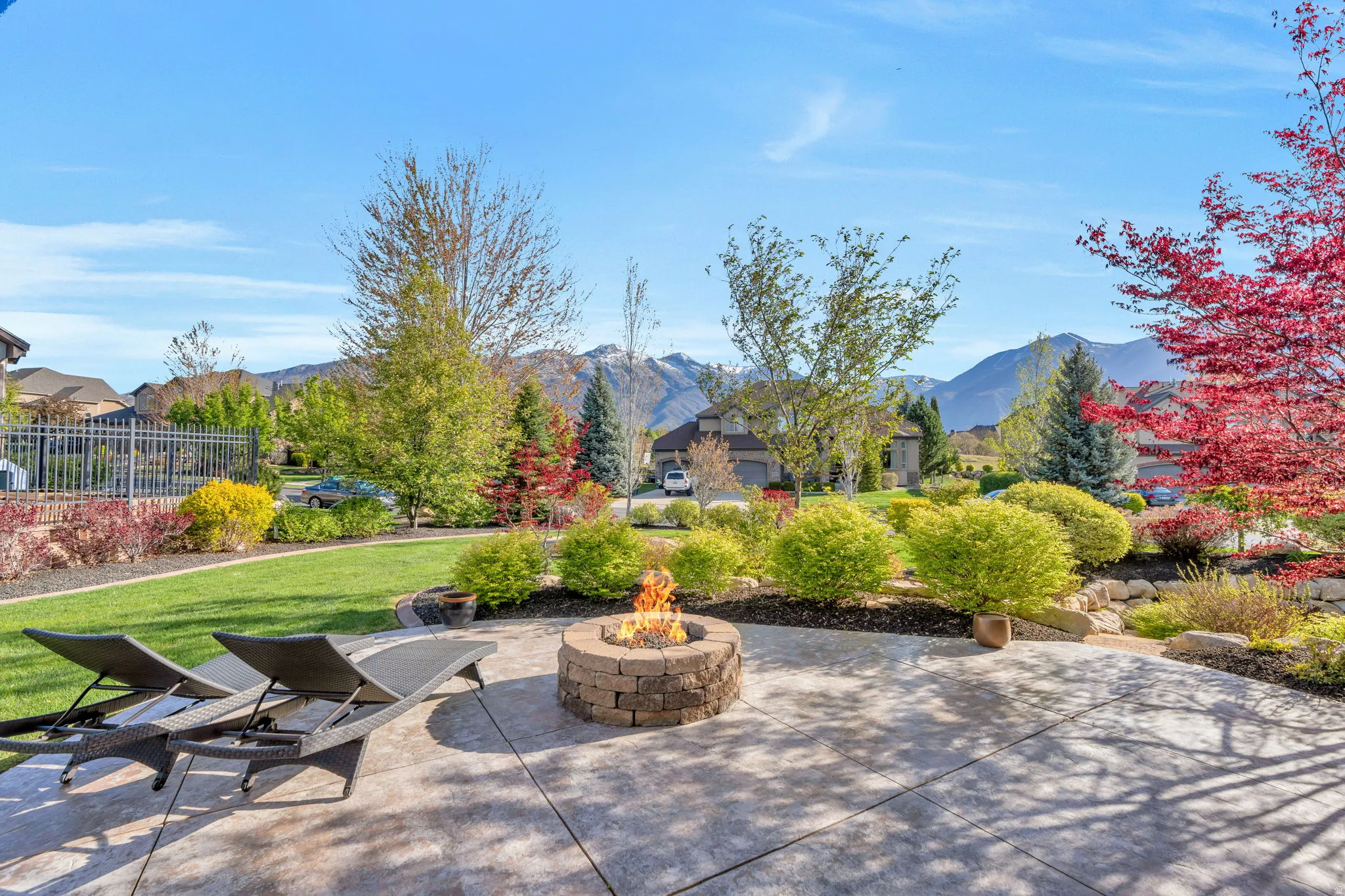 Spacious concrete patio featuring a built-in stone fire pit, surrounded by lush landscaping, a manicured lawn, and distant mountains