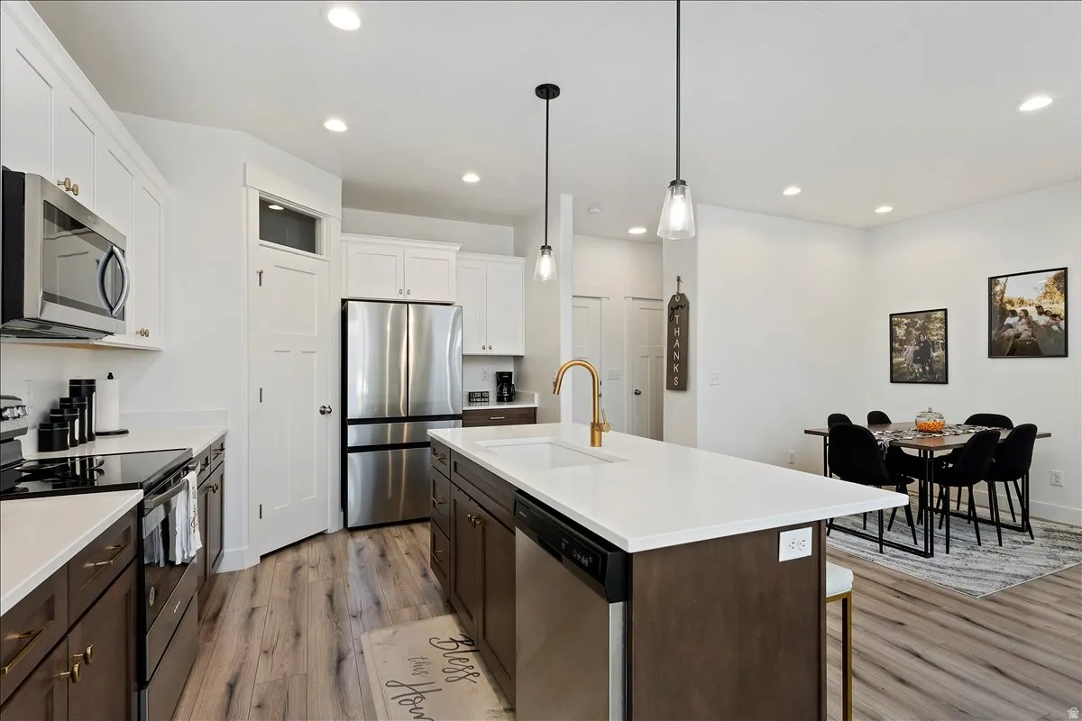 Dual tone kitchen with stainless steel appliances, two tone cabinetry, light wood-style floors, an island with sink, and decorative light fixtures