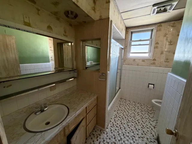 Full bath featuring wallpapered walls, vanity, combined bath / shower with glass door, tile walls, and wainscoting