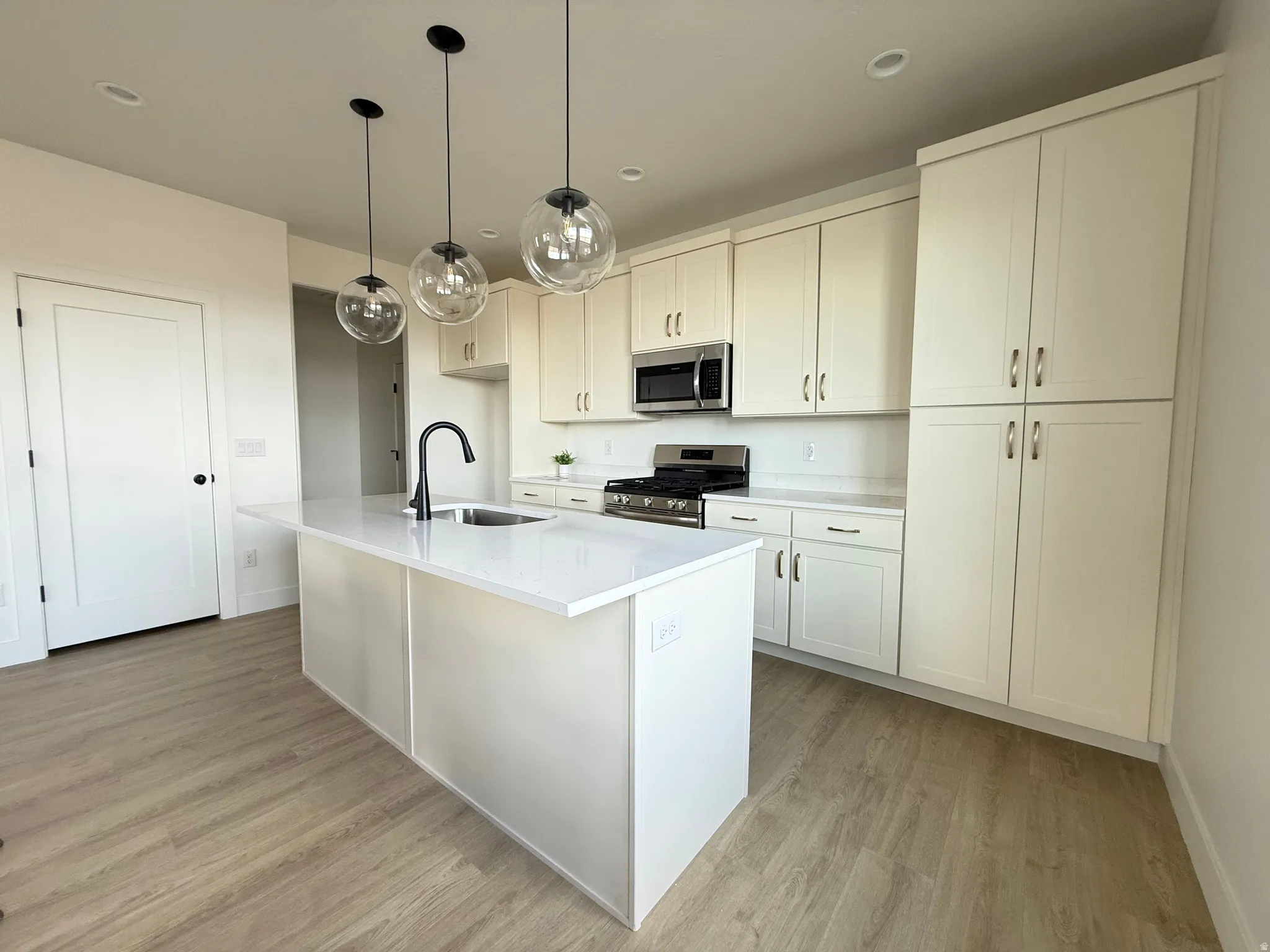 Kitchen featuring an island with sink, stainless steel appliances, light wood-style floors, hanging light fixtures, and white cabinetry