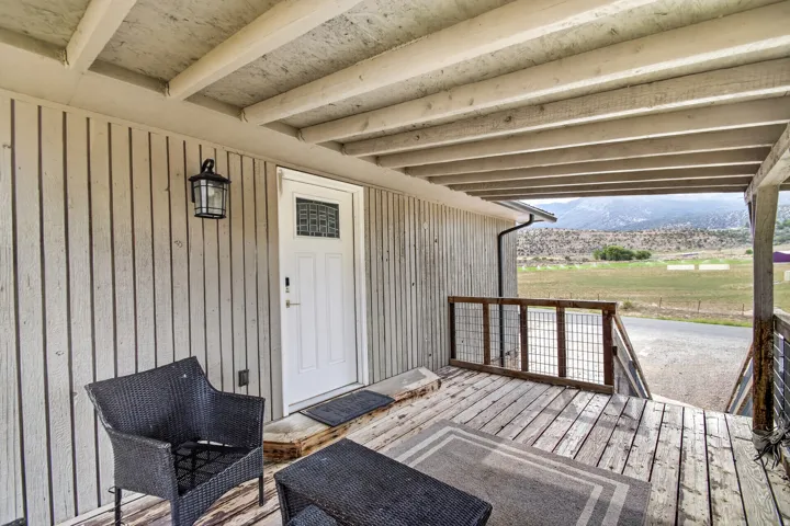 Unit 1: Covered deck with wood-finish flooring and exposed beam ceiling, featuring a white entry door, black wicker seating, and metal railing