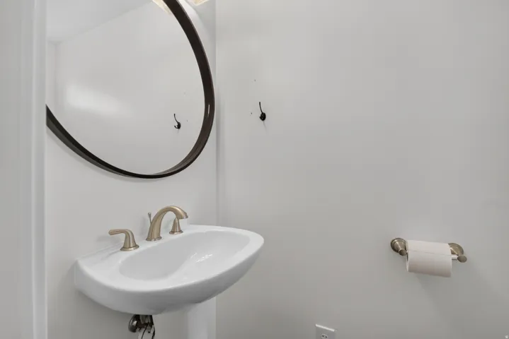 Unit 2B: Pedestal sink with brushed metal faucet, oval mirror with dark frame, and wall-mounted toilet paper holder
