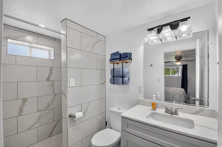 Unit 2A: Walk-in shower with tiled walls and a transom window, accompanied by a vanity featuring a rectangular undermount sink, a light-toned countertop, and a wide mirror