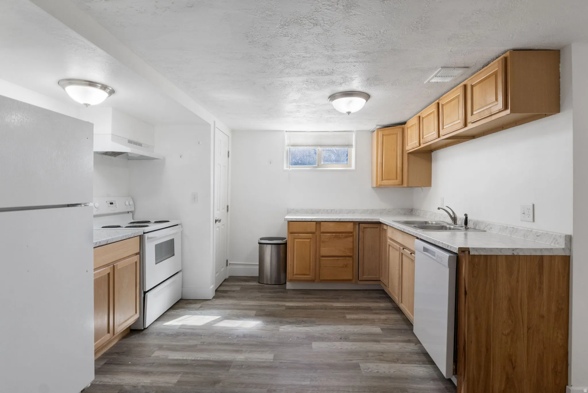 Kitchen with white appliances, a textured ceiling, light countertops, dark vinyl flooring, and light wood finish cabinets