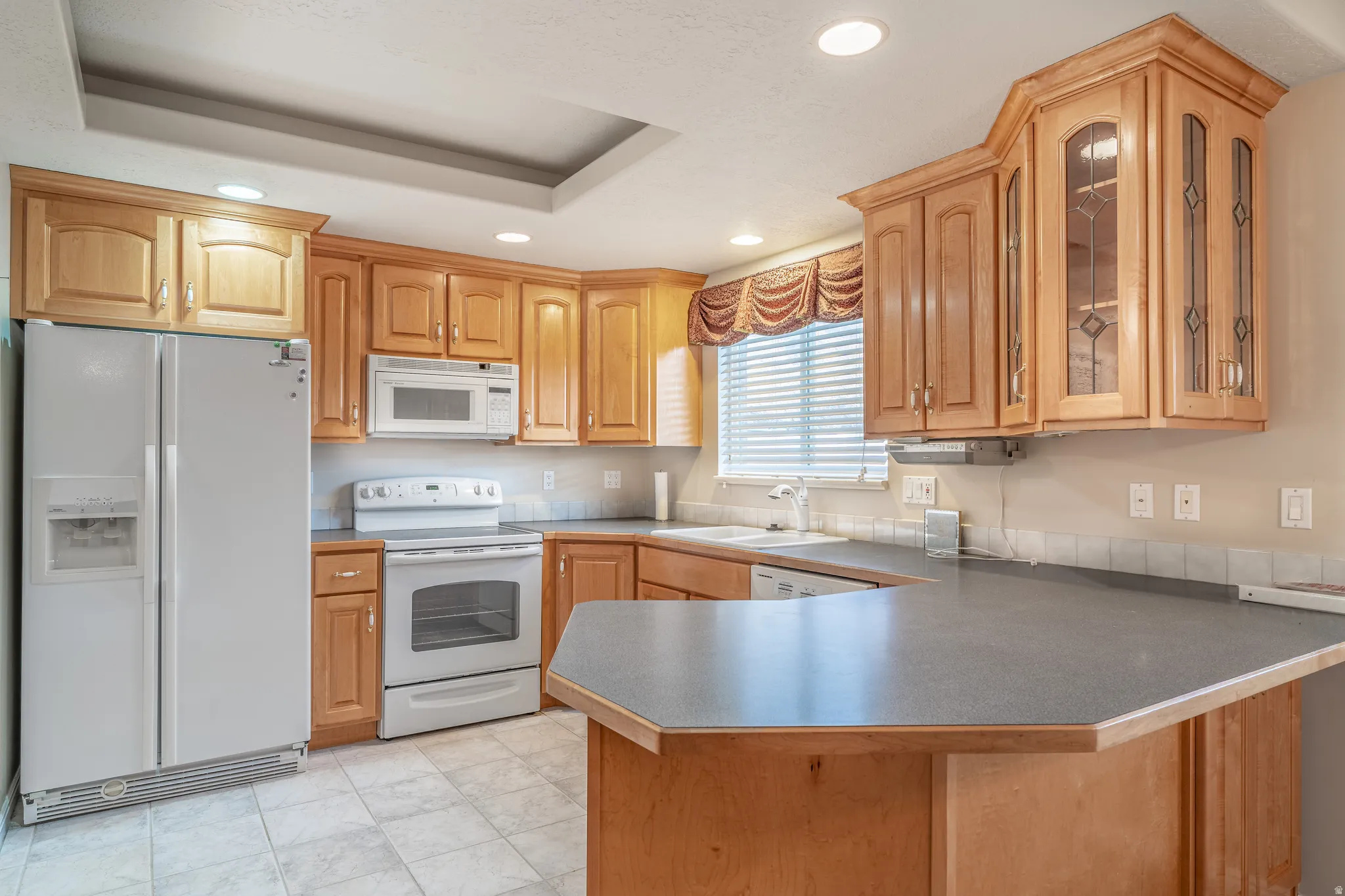 Kitchen with white appliances, a peninsula, recessed lighting, glass fronted cabinets, and a tray ceiling