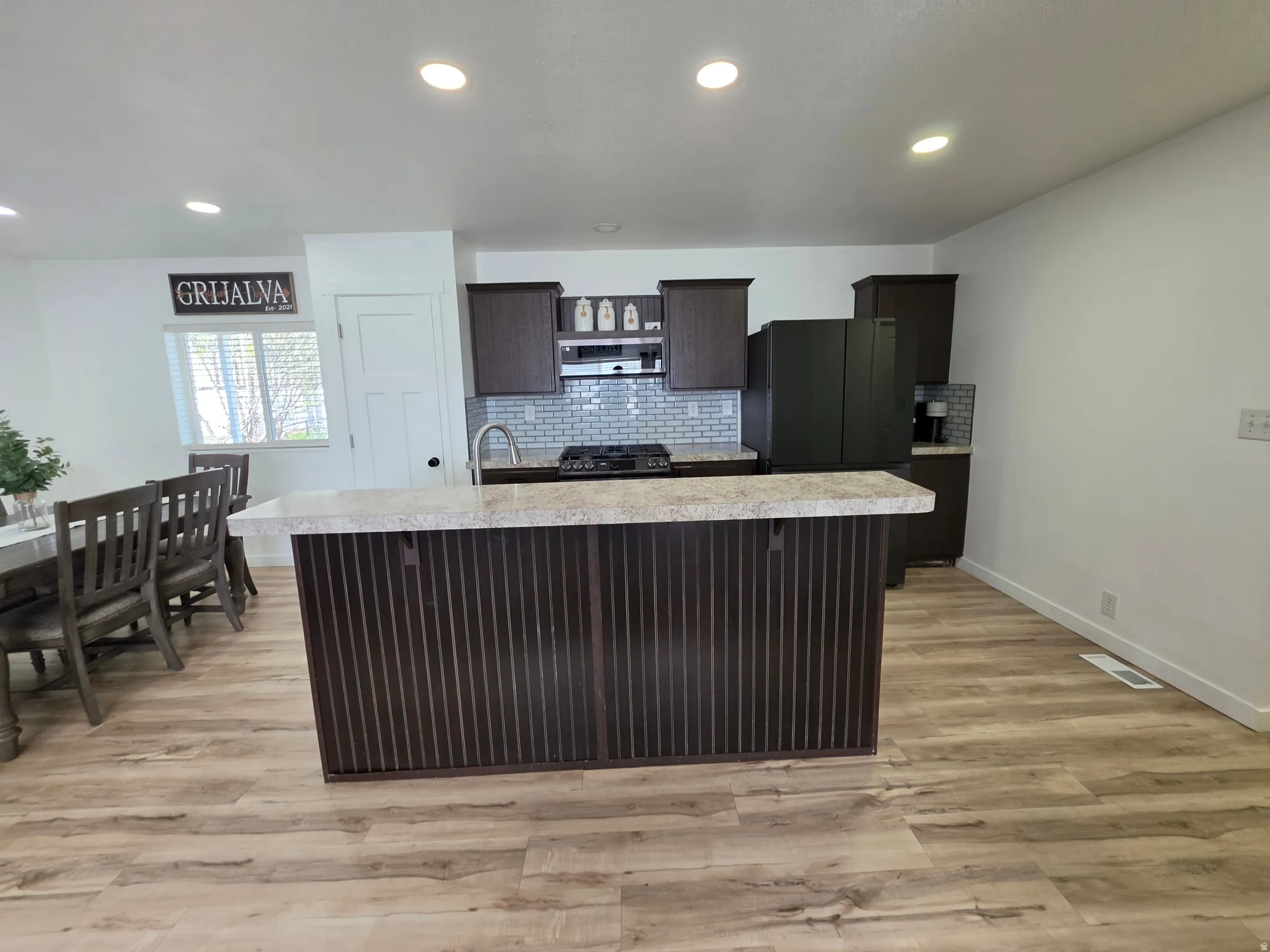 Kitchen featuring dark wood finish cabinetry, light countertops, a center island with sink, light wood-style floors, and tasteful backsplash