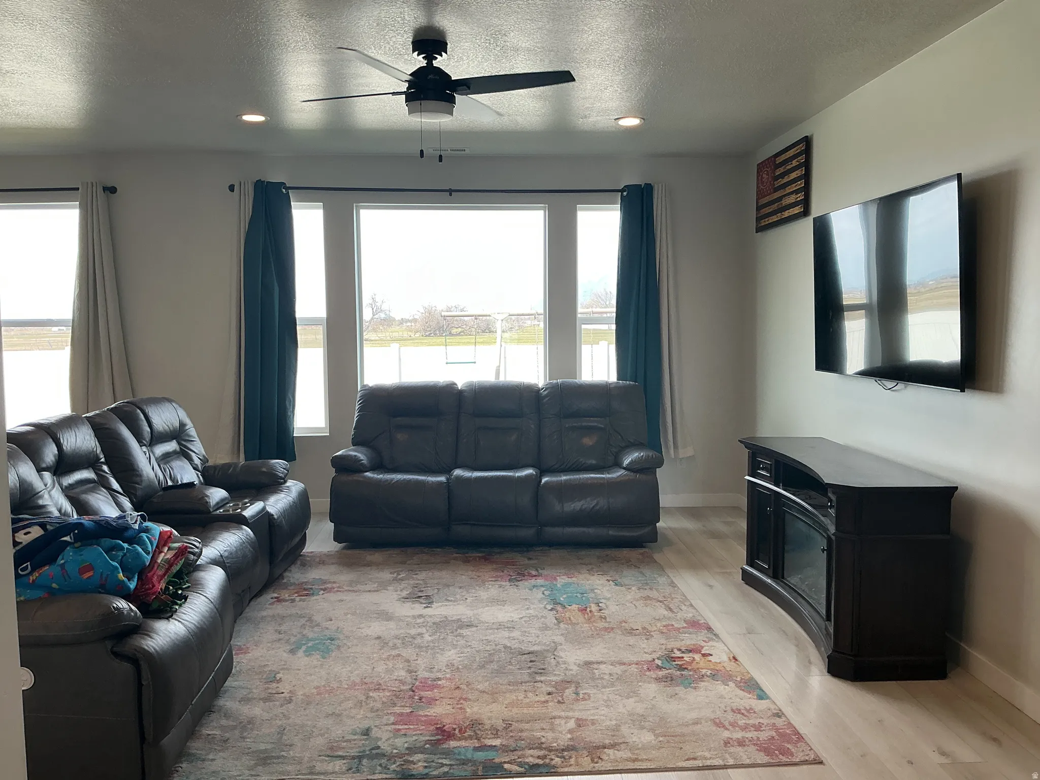 Living room with a ceiling fan, light wood-type flooring, a wood stove, a textured ceiling, and plenty of natural light
